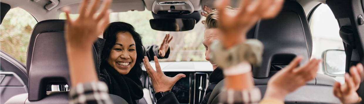 Två vuxna sitter i framsätet på en bil och ler, medan två barn i baksätet höjer armarna i luften och ser glada ut under bilresan.