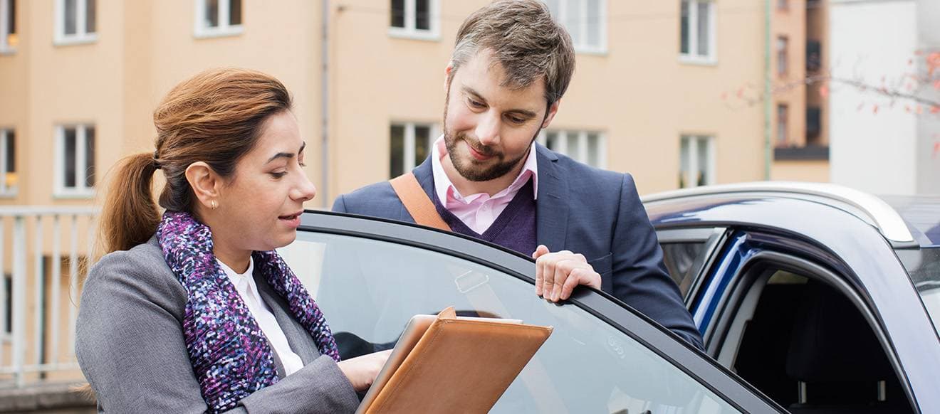 Två personer vid en bil, varav den ena håller upp papper som de tittar på tillsammans.