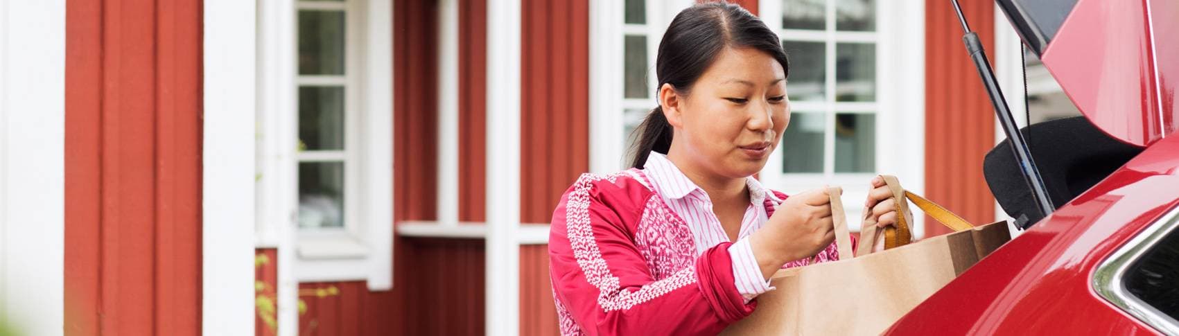Kvinna som bär ut matkasse ur bagagelucka framför ett hus