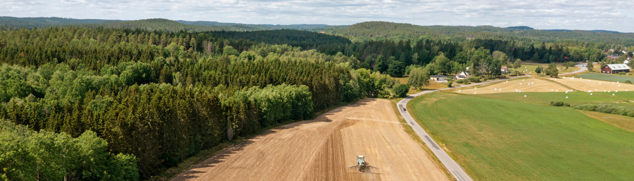 Flygbild över jordbrukslandskap med åkrar, skog och en traktor på ett fält.