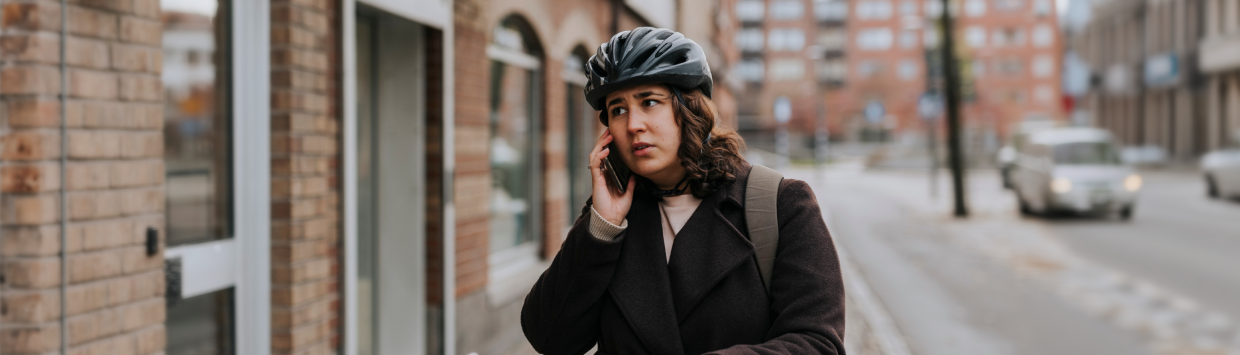En kvinna med cykelhjälm står på en trottoar och pratar i mobiltelefon.
