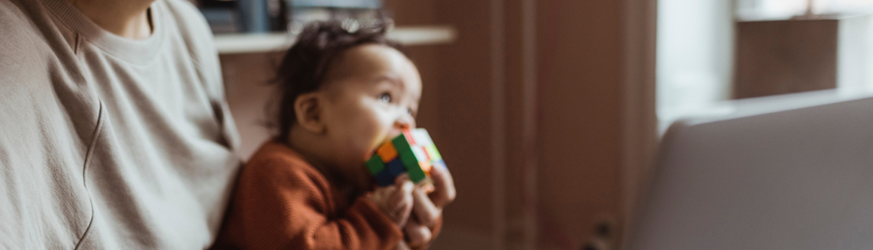 En vuxen person sitter vid en dator med en bebis i famnen. Bebisen har en Rubiks kub delvis i munnen.